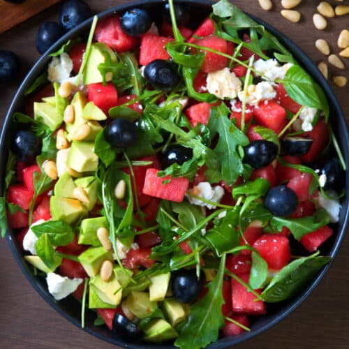 Top view of watermelon salad in a black bowl, pine nuts and blueberries on the side.