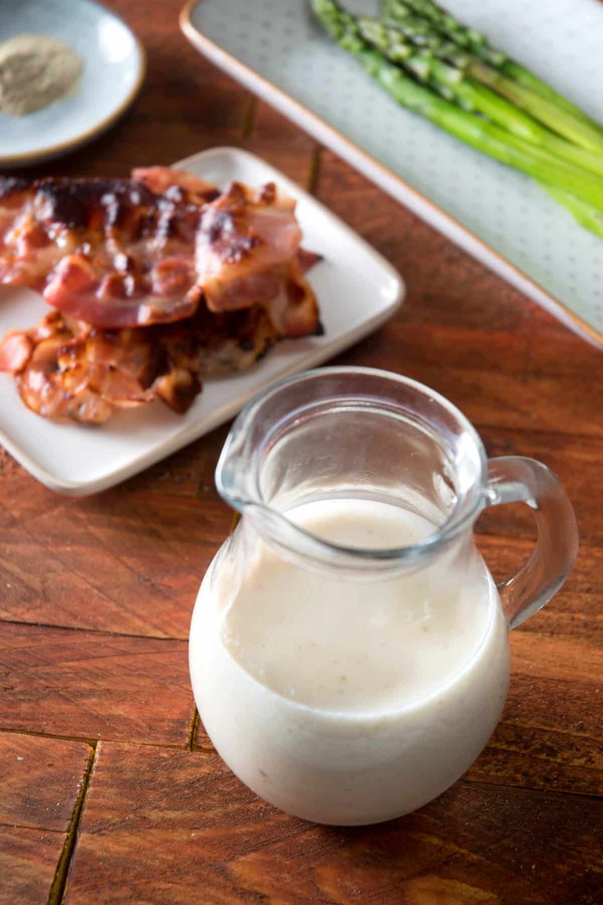 White gravy in a jar with bacon and asparagus in the background.