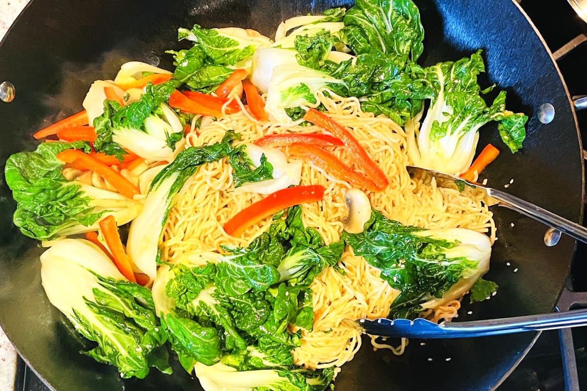 Bok choy stir fry with noodles and tongs in wok.