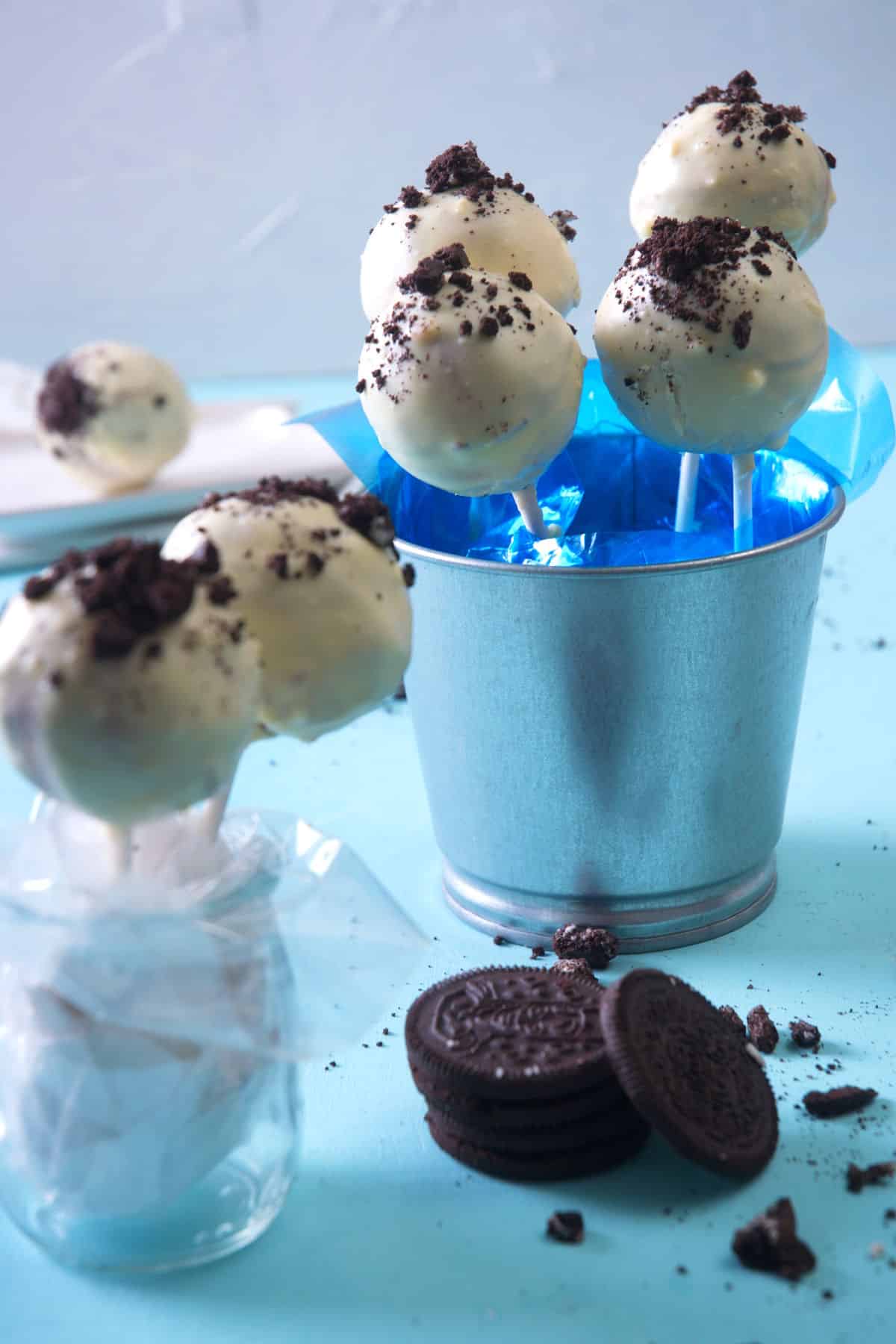 Cookies and cream cake pops in a decorative, blue, mini pail with Oreo cookies on the side.