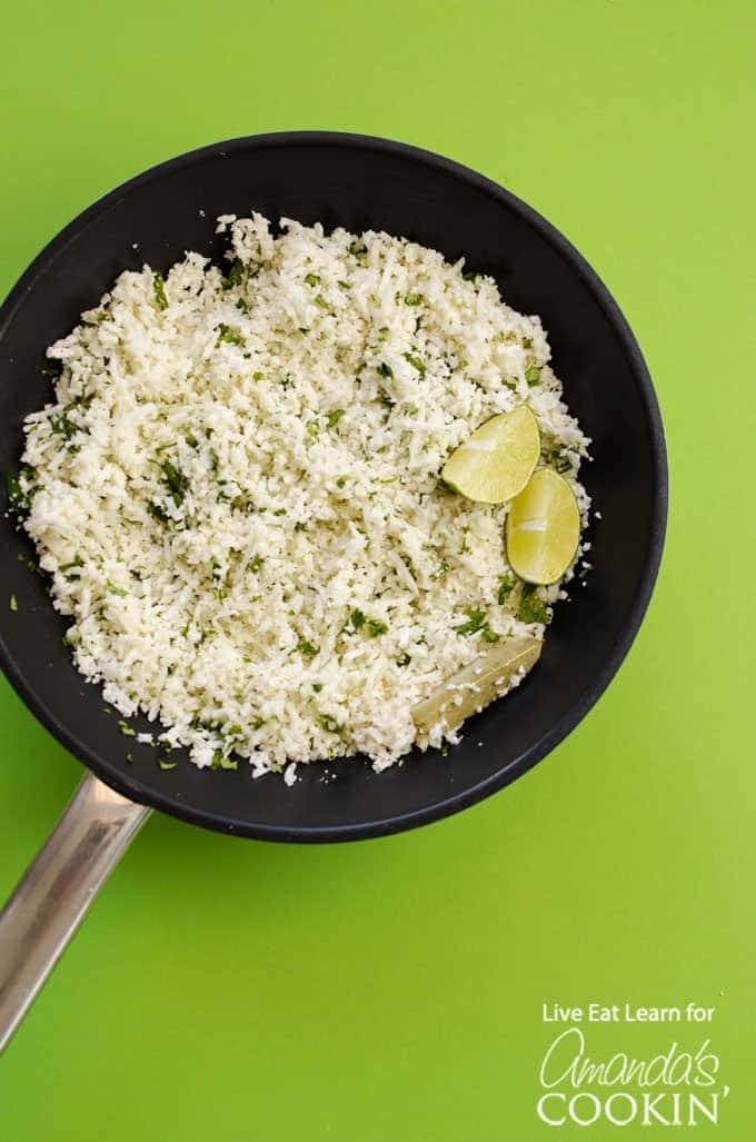 Cauliflower rice in pan with cilantro and lime on green background.