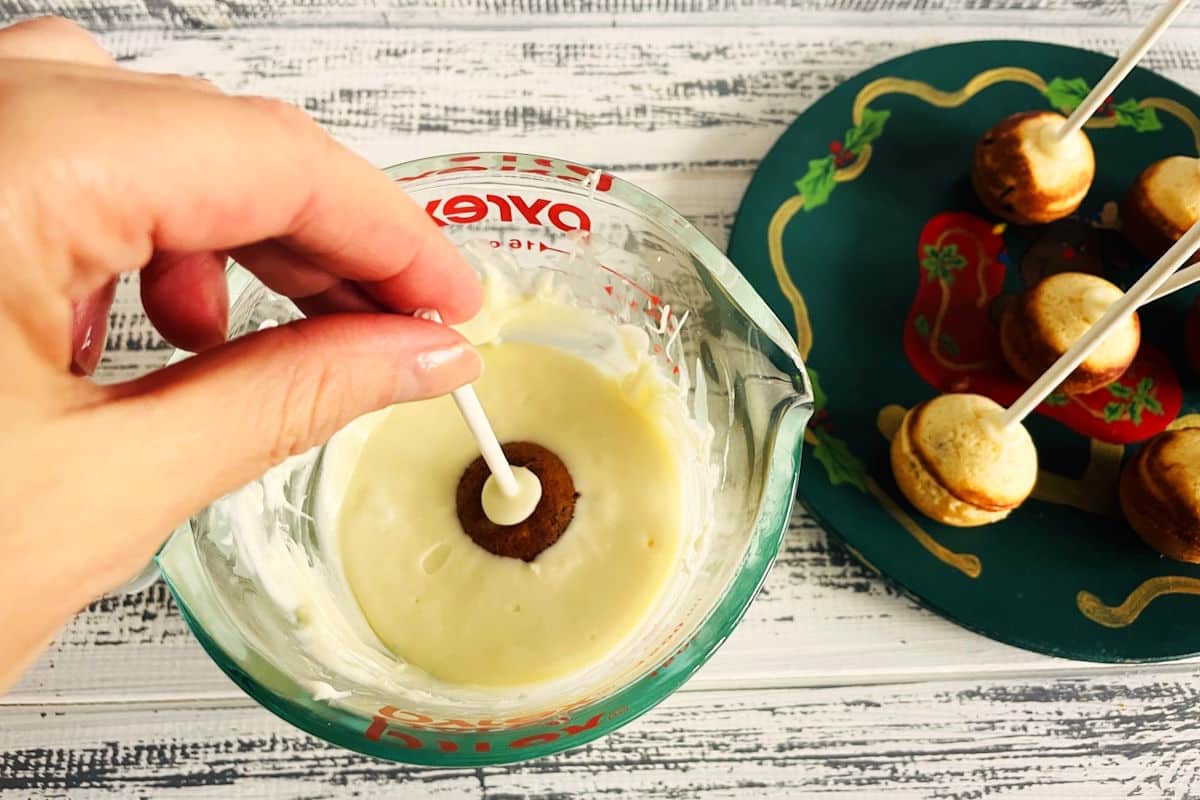 Cranberry cake pop getting dipped into white chocolate on white wooden background.
