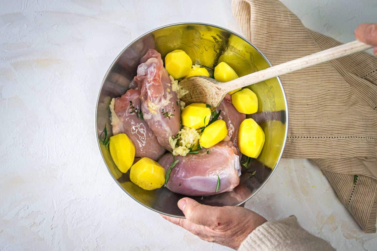 Chicken thighs, herbs and baby potatoes in bowl with wooden spoon.
