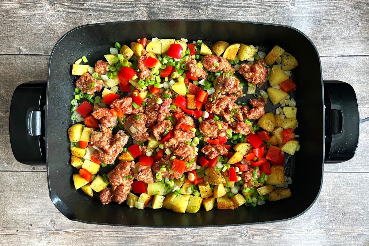 Breakfast hash ingredients frying in electric skillet.
