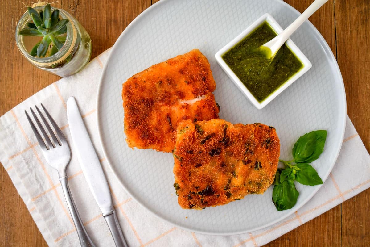 Electric skillet fried fish on white plate with fresh basil, pesto sauce and cutlery on the side.