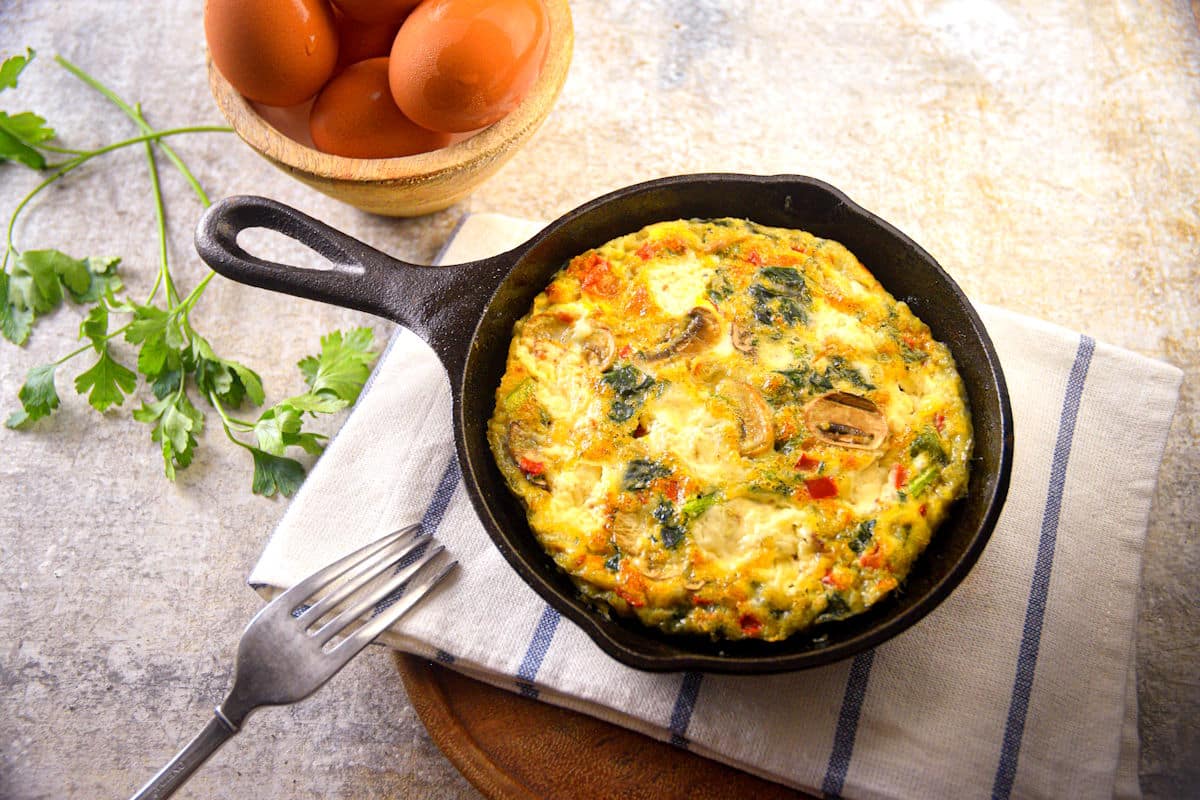 Electric skillet frittata in pan on kitchen towel with fork and eggs on the side.