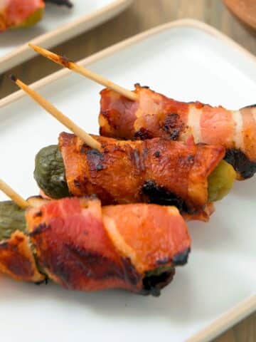 Grilled bacon-wrapped pickles on toothpicks on white dish.