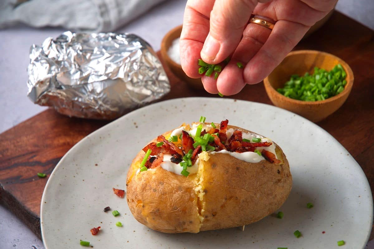 Baked potato with sour cream, chives and bacon bits on plate with woman's hand.