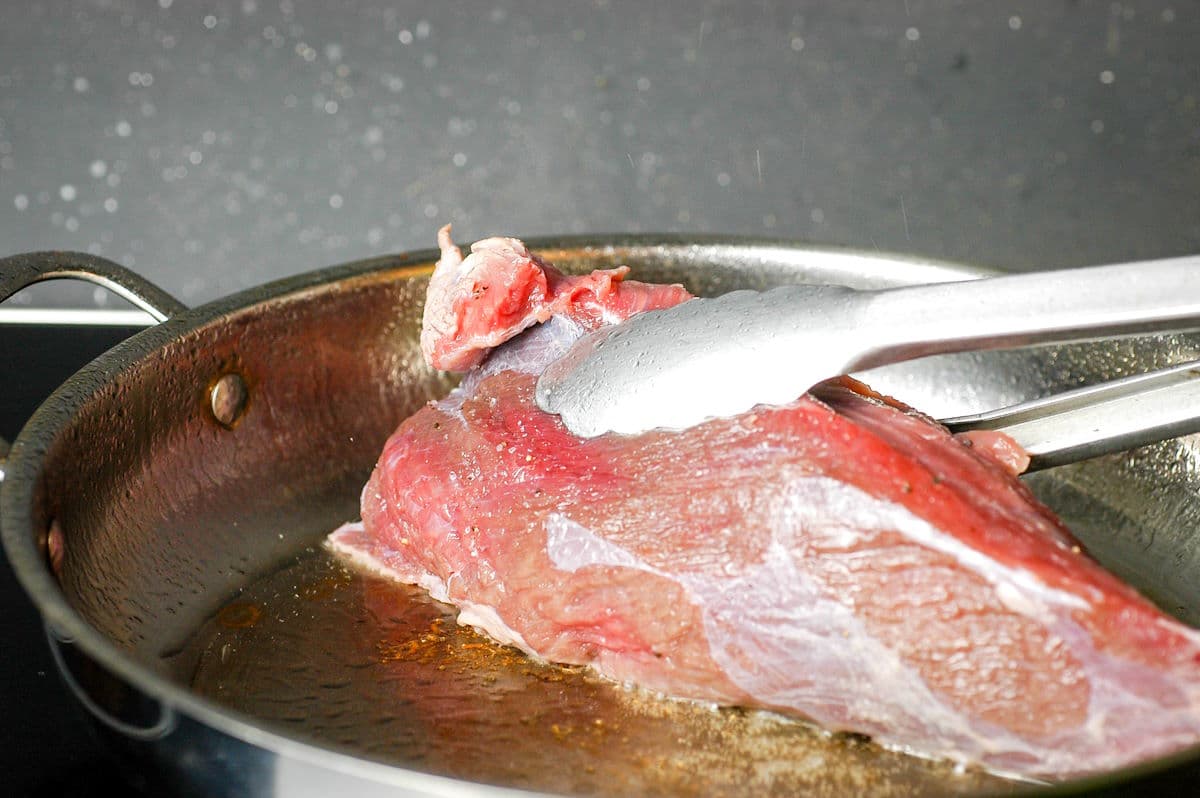 Beef roast searing in pan with tongs.