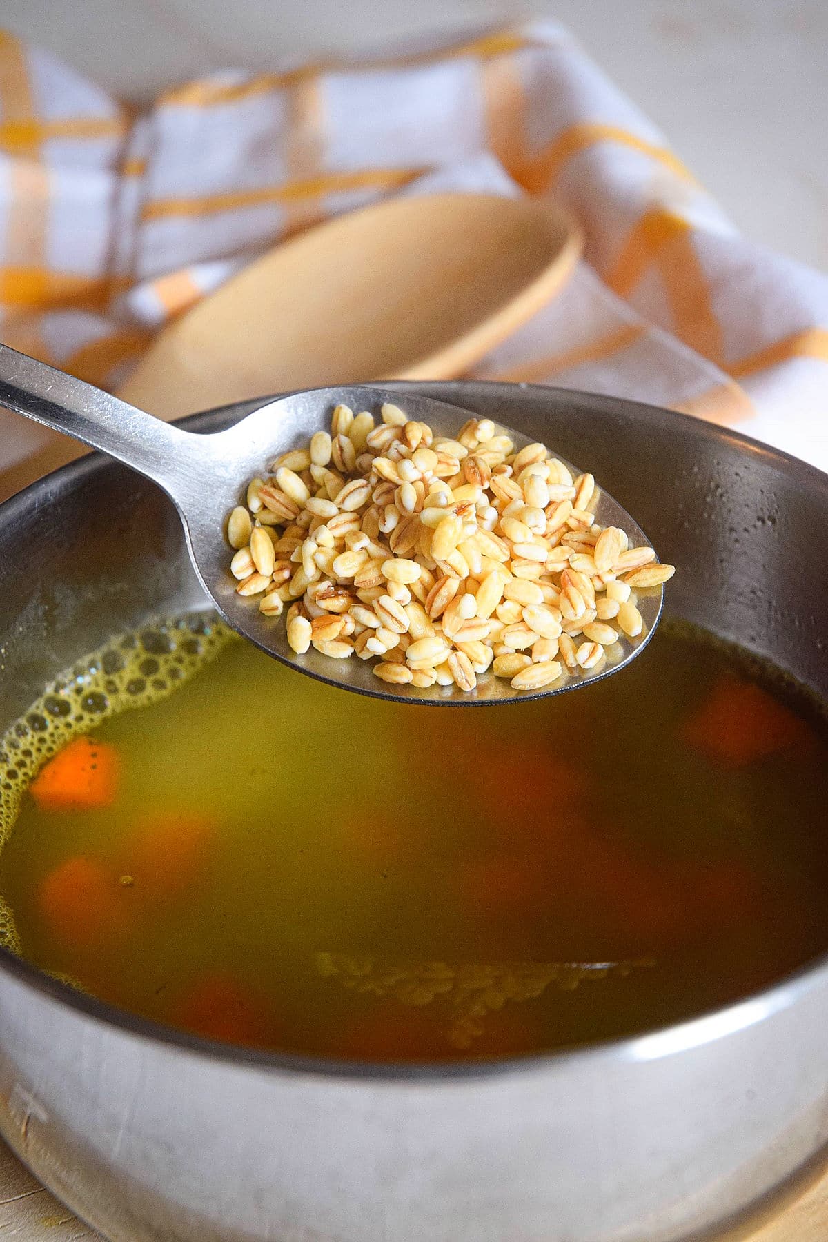 Uncooked pearl barley in spoon over a pot of soup.