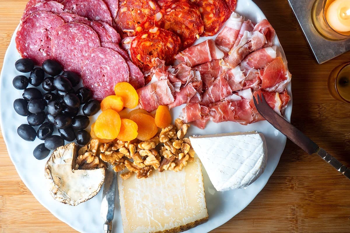 Circular charcuterie board with meat, cheese, breadsticks, nuts, apricots and olives.