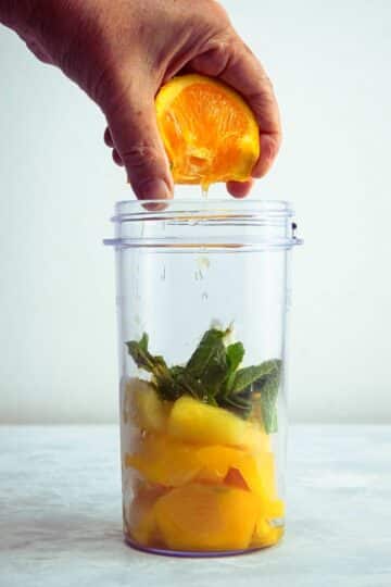 Mango pineapple smoothie ingredients in blender jar with orange being squeezed over top.