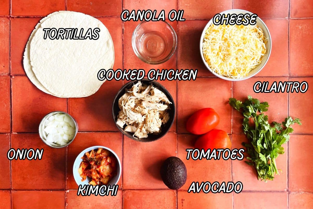 Kimchi quesadilla ingredients labeled on terra cotta tile background.