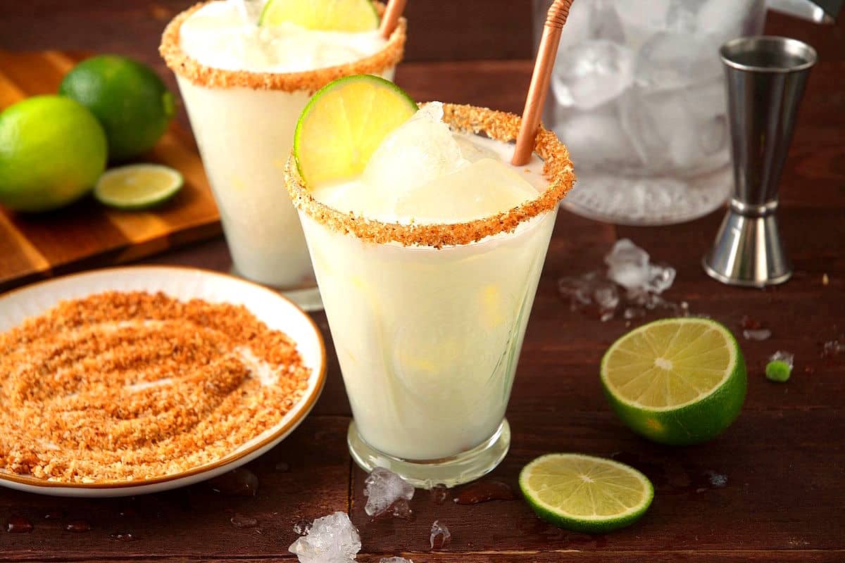 Lime cremosa drink in glasses with straws, lime slices and toasted coconut on the side.