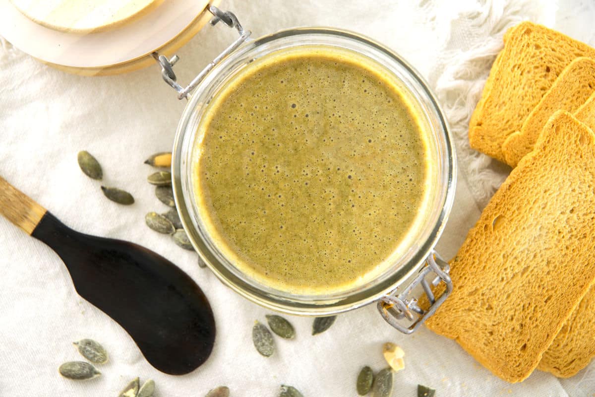 Pumpkin seed butter in jar with crackers, spatula and pumpkin seeds on the side.