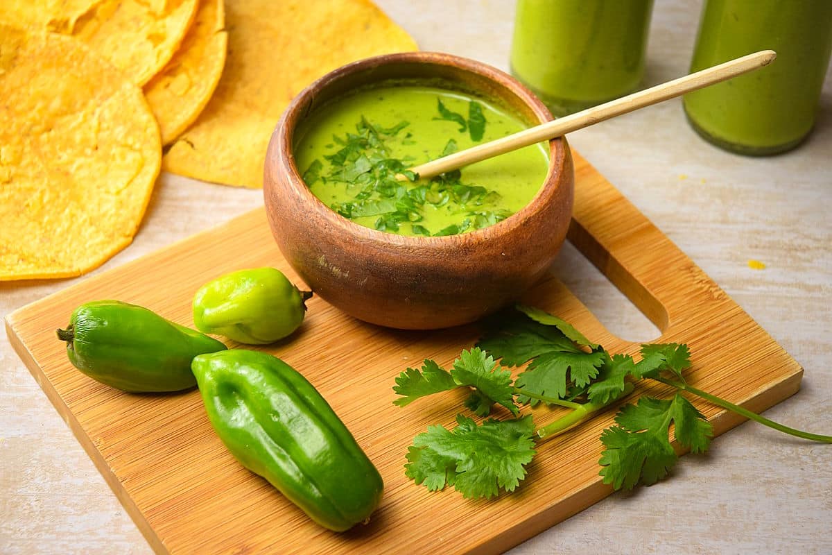 Tomatillo hot sauce in bowl with cilantro, tortilla chips and jalapenos on the side.